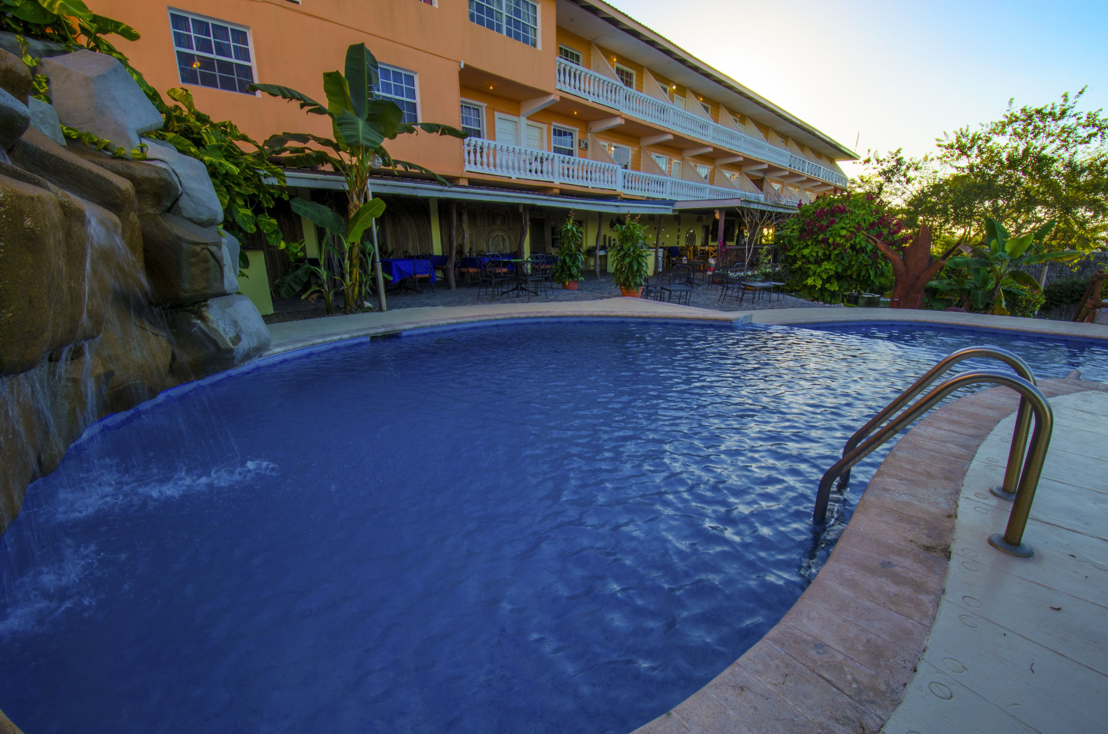 A swimming pool with a stone waterfall feature, surrounded by a building with balconies and outdoor seating.