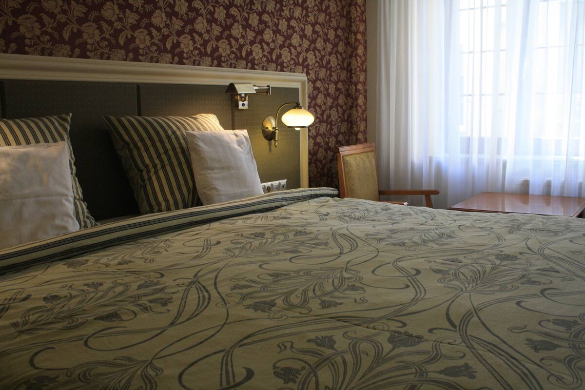 A hotel room with a large bed, a nightstand, a lamp, and a view of the outside through a window.