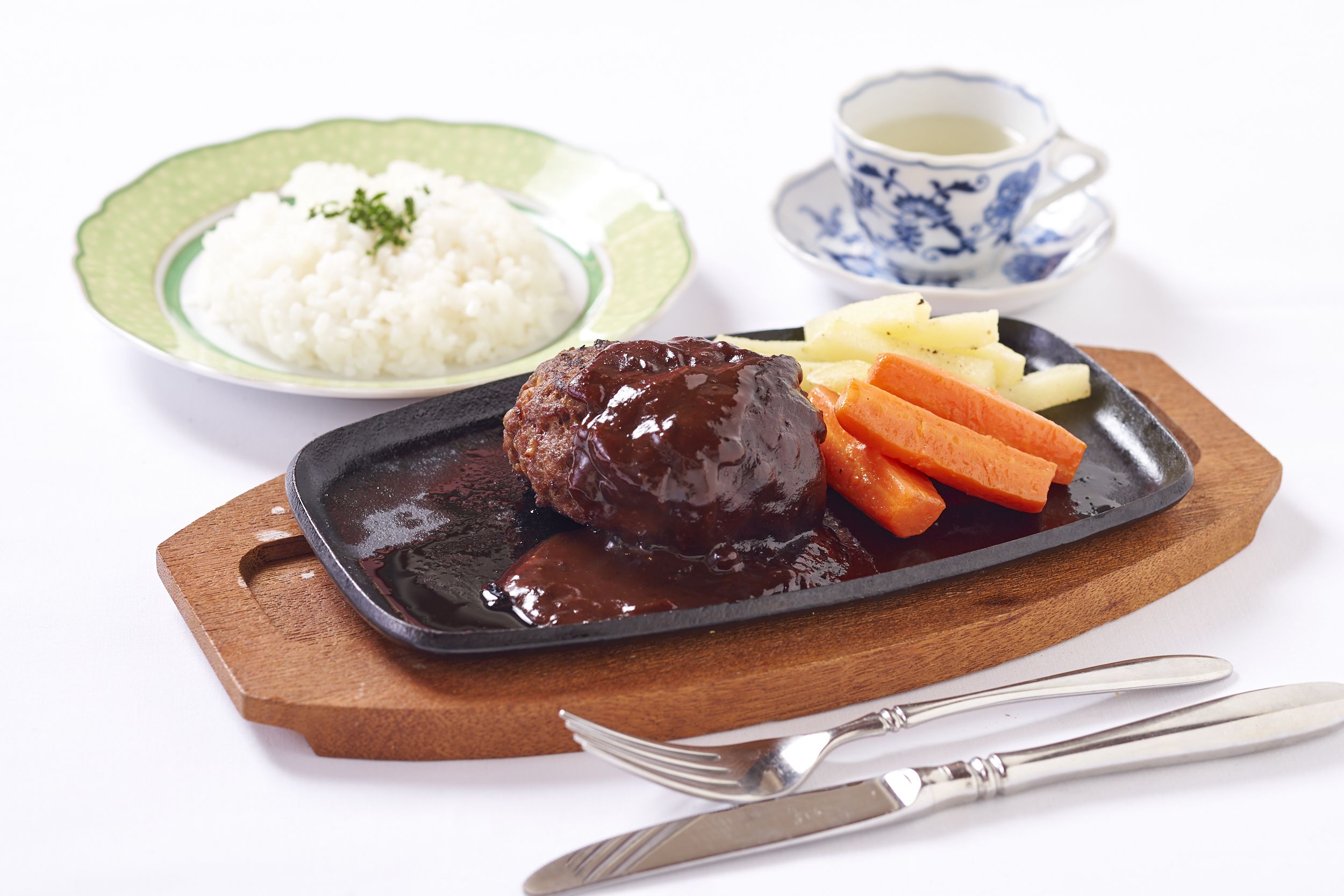 Un plato con carne, arroz y verduras sobre una tabla de madera, acompañado de una taza y un tenedor.