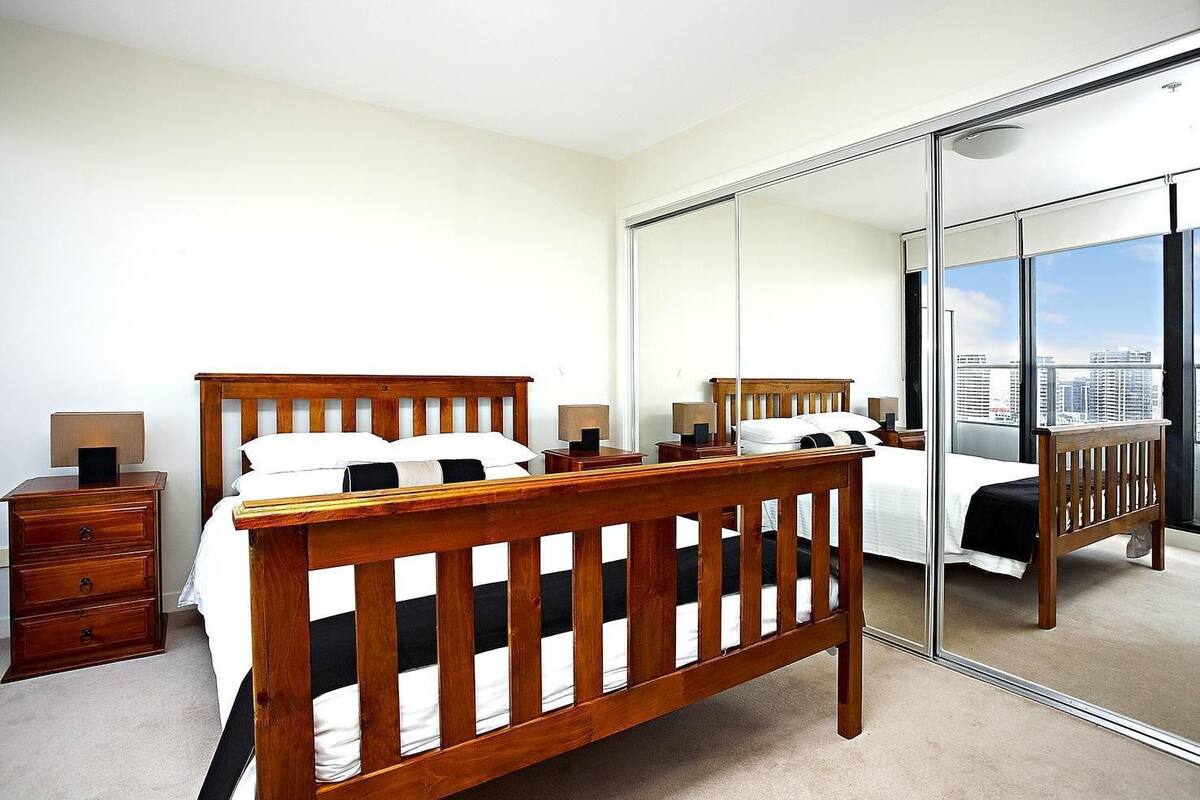 Un dormitorio con una cama de madera, una mesita de noche, un aparador y un espejo grande que refleja una vista de la ciudad.