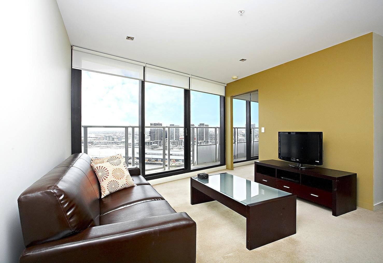 Un salon moderne avec un canapé en cuir marron, une table basse en verre et un téléviseur à écran plat posé sur un meuble en bois. De grandes fenêtres offrent une vue sur la ville.