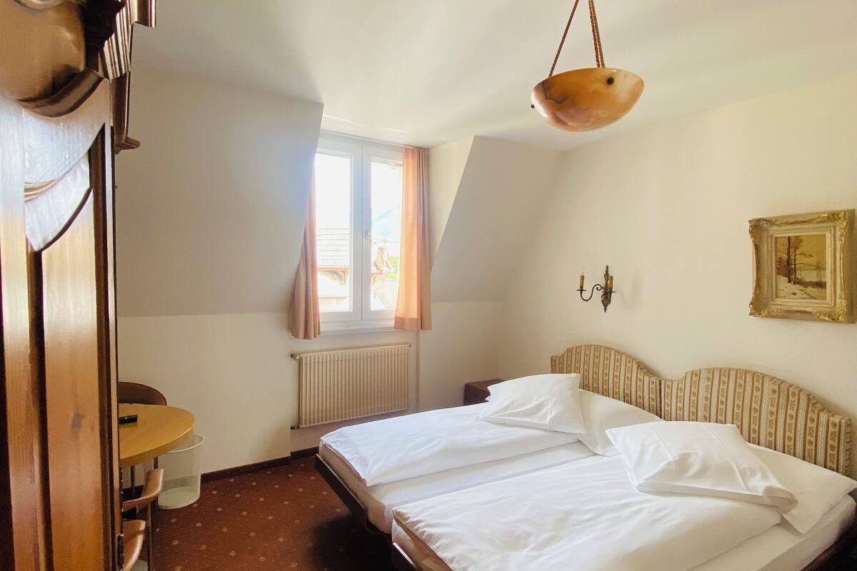 Une chambre d’hôtel avec un lit double, une armoire en bois, une petite table avec des chaises, un radiateur et une fenêtre avec des rideaux.
