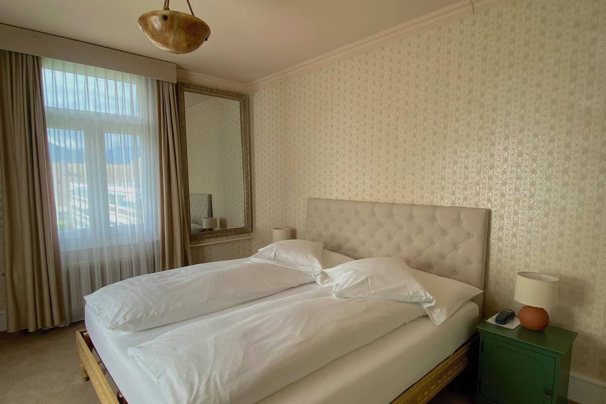 Un lit double bien fait, avec du linge de lit blanc, une table de chevet verte avec une lampe, et une grande fenêtre avec des rideaux.