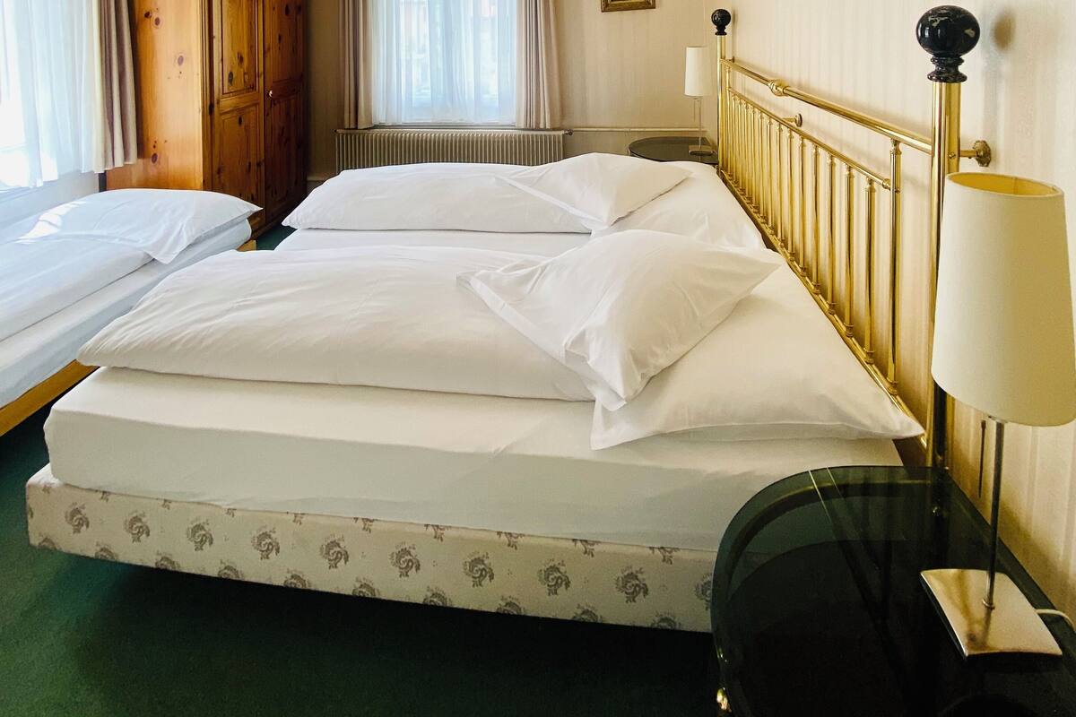 Une chambre d’hôtel avec deux lits, une armoire en bois, un radiateur, une lampe et une fenêtre avec des rideaux.