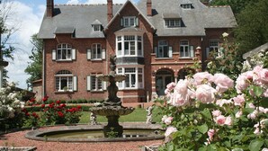 Un edificio de ladrillo grande con una torre central, una fuente y un jardín de rosas rosadas.