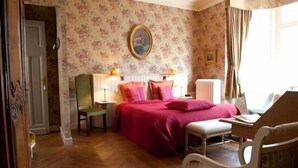 Un dormitorio con papel tapiz floral, una cama con ropa de cama rosa, suelo de madera, una silla, una mesita de noche y una ventana con cortinas.
