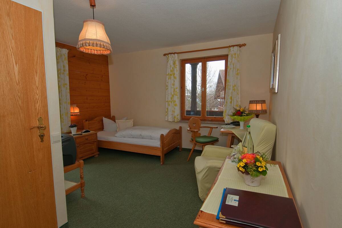 A room with a bed, a desk with a chair, a table with a vase of flowers, and a window with curtains.