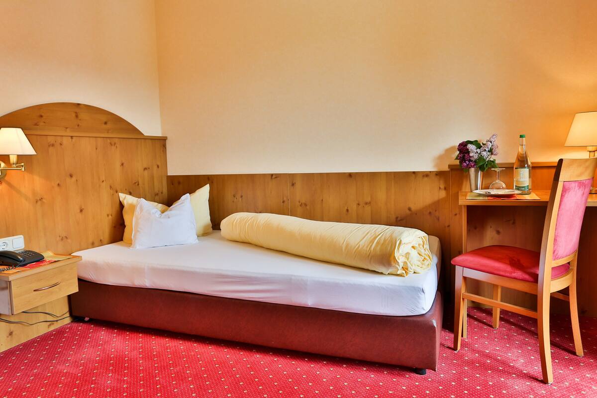 A hotel room with a wooden headboard, a single bed with white bedding, a red polka dot carpet, a wooden nightstand with a lamp, a wooden chair, and a small table with a vase of flowers and a bottle of wine.