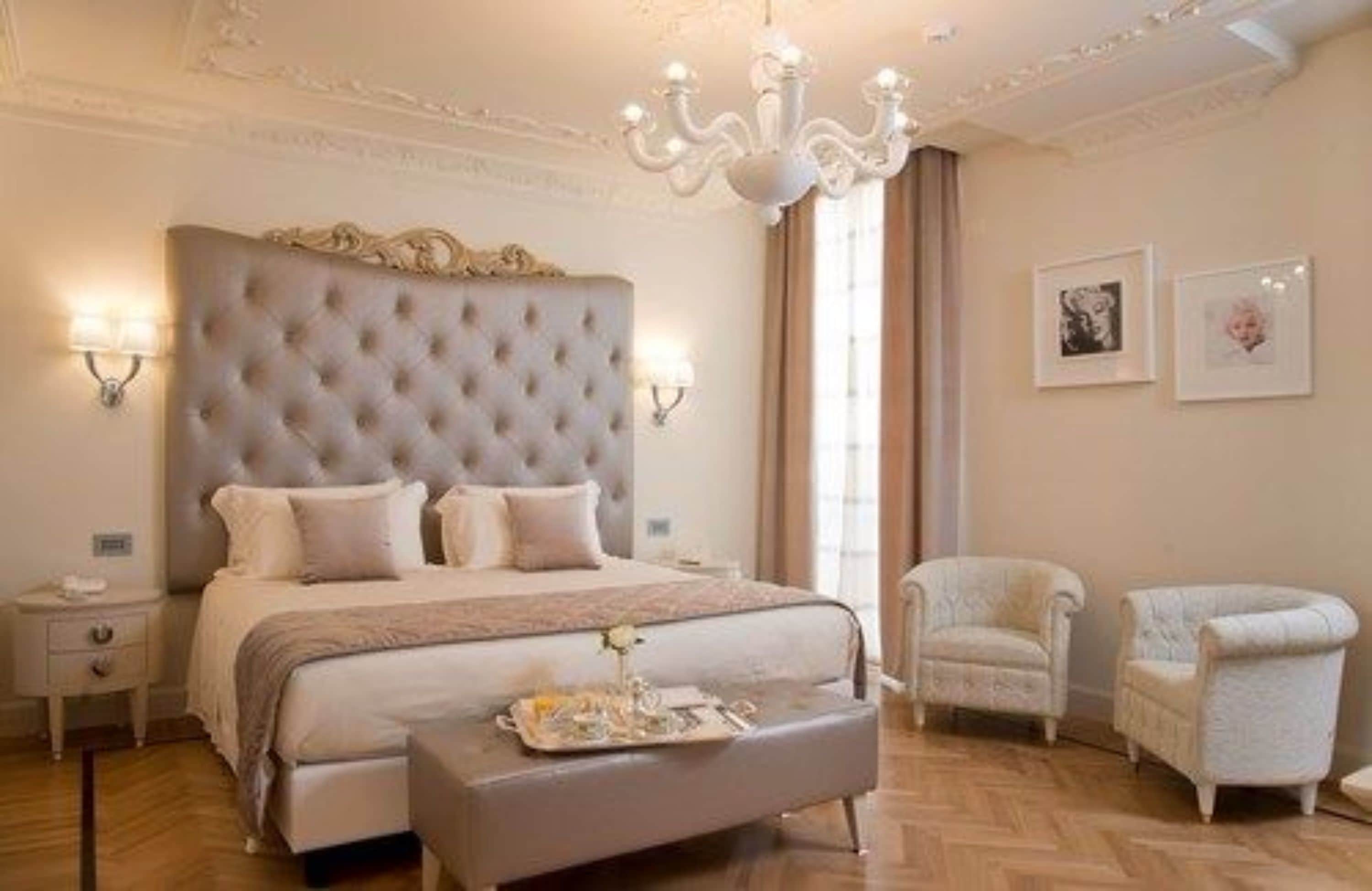A bedroom with a tufted headboard, a bedside table, a nightstand, a bench with a tray, two armchairs, and a chandelier.