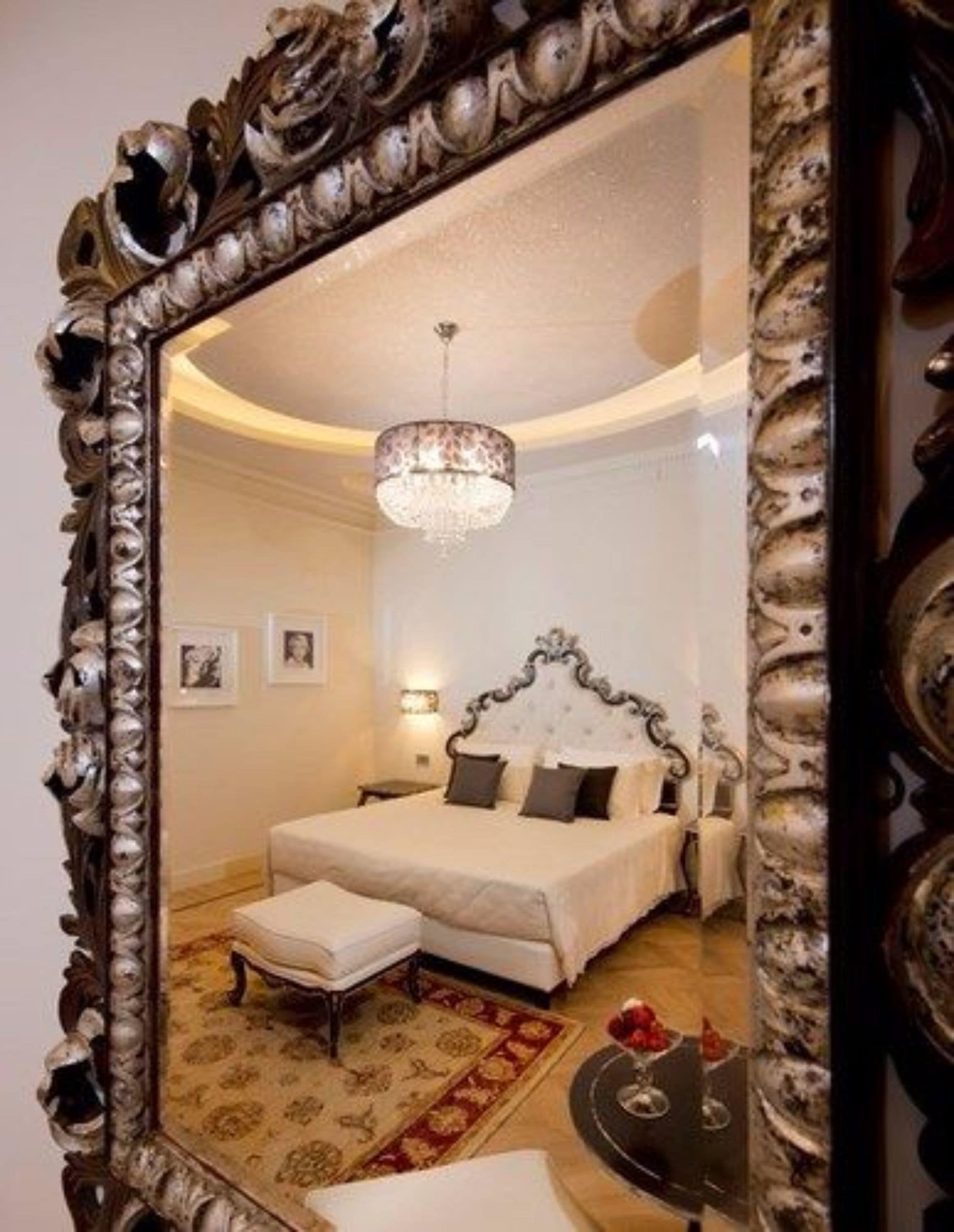 Un dormitorio con un espejo grande y ornamentado, una araña, una cama con sábanas blancas y una mesita con un objeto rojo.