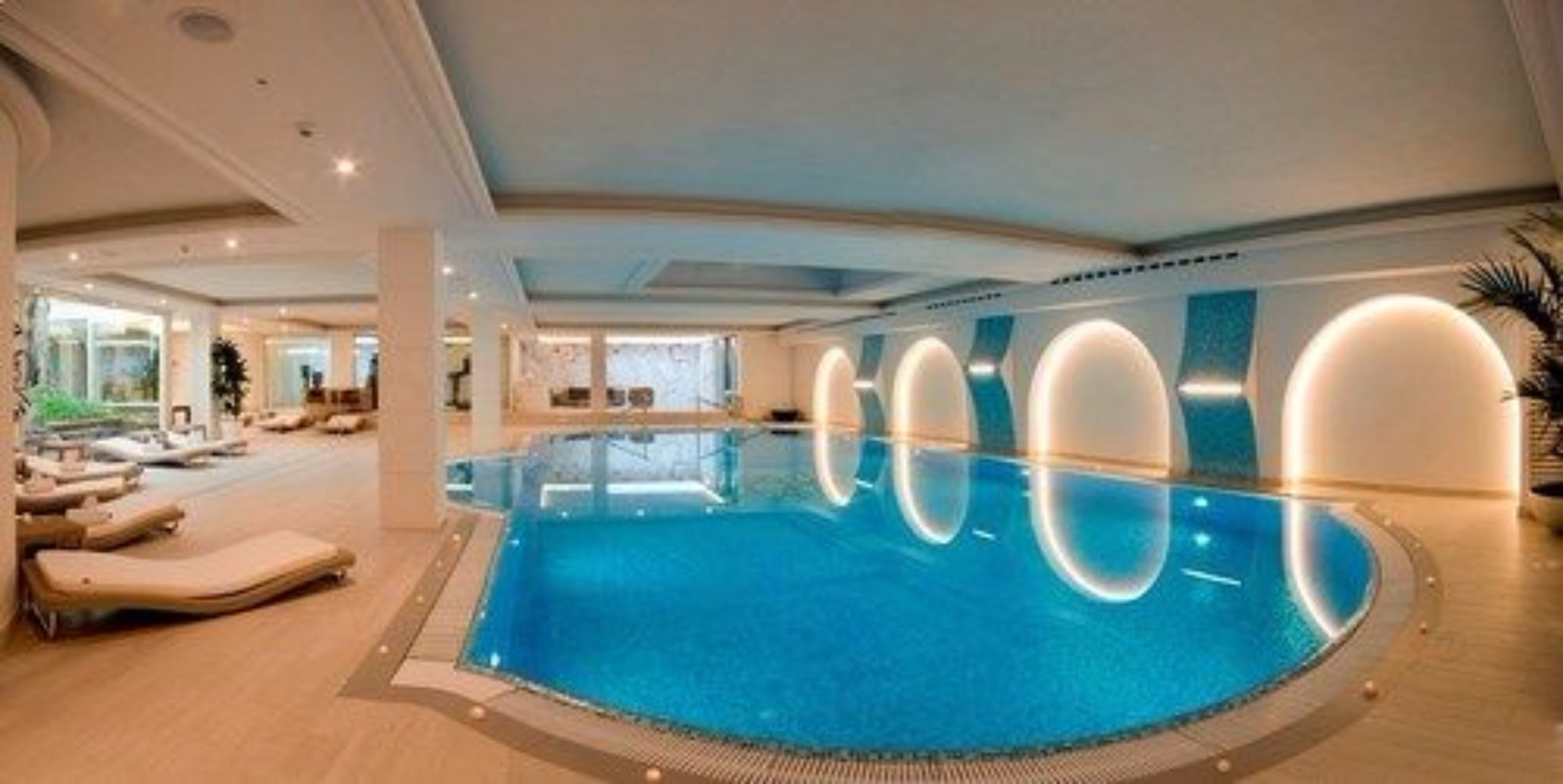 Une grande piscine intérieure au design moderne, entourée de chaises longues et dotée d’un plafond bien éclairé.