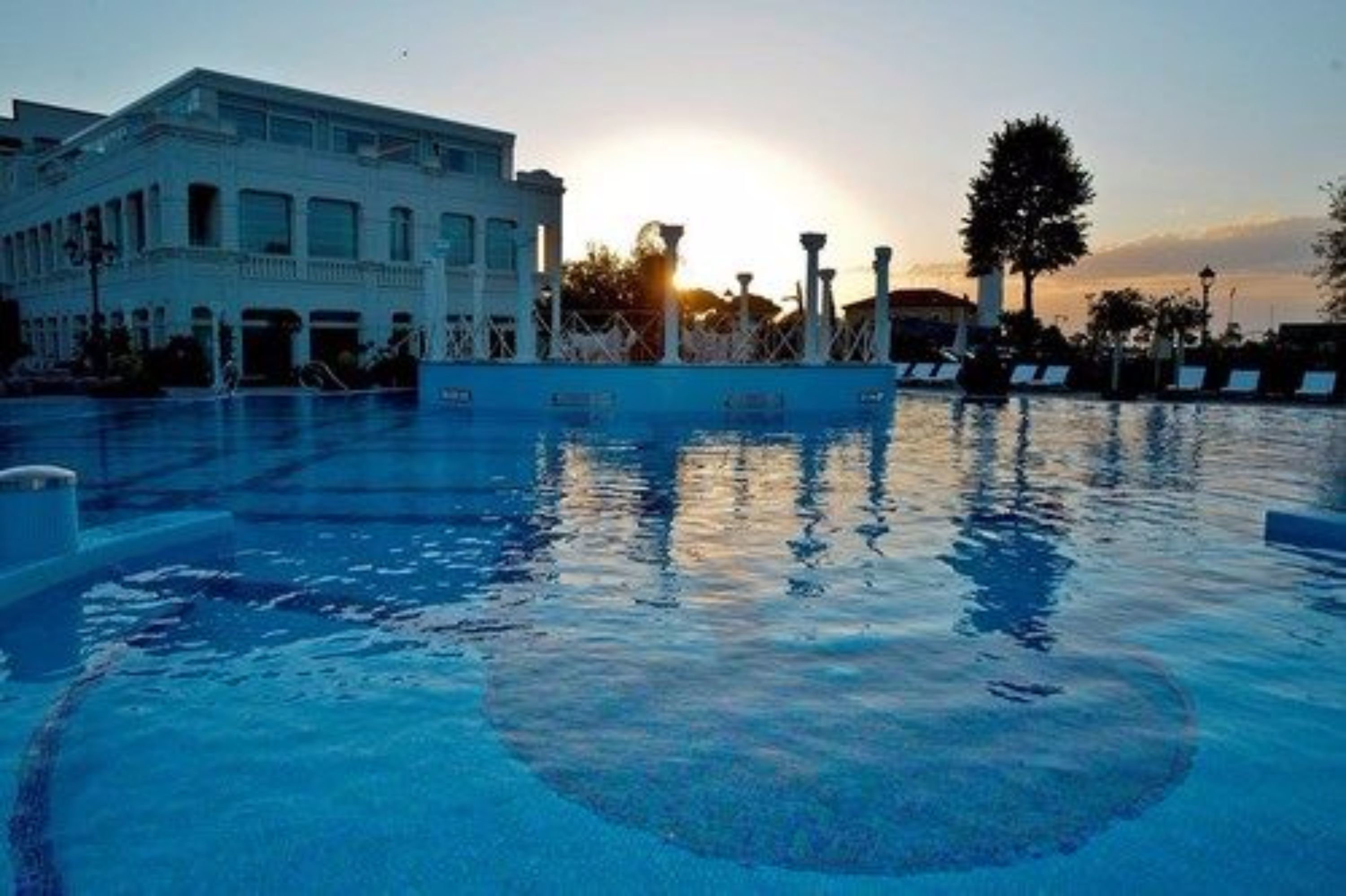Una piscina con un atardecer al fondo, un edificio con balcón y un árbol.