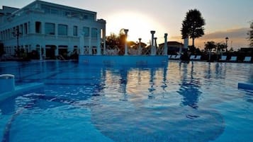 Una piscina con un atardecer al fondo, un edificio con balcón y un árbol.