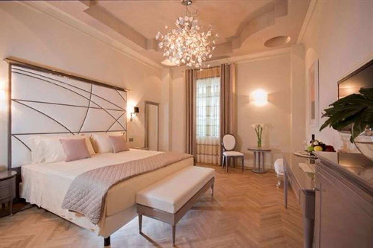 A bedroom with a large bed, a bench, a desk, a chair, a chandelier, and a mirror.