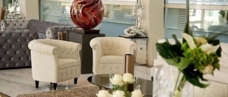 A modern living room with a grey tufted sofa, two cream-colored armchairs, a glass coffee table, and a decorative glass sculpture.