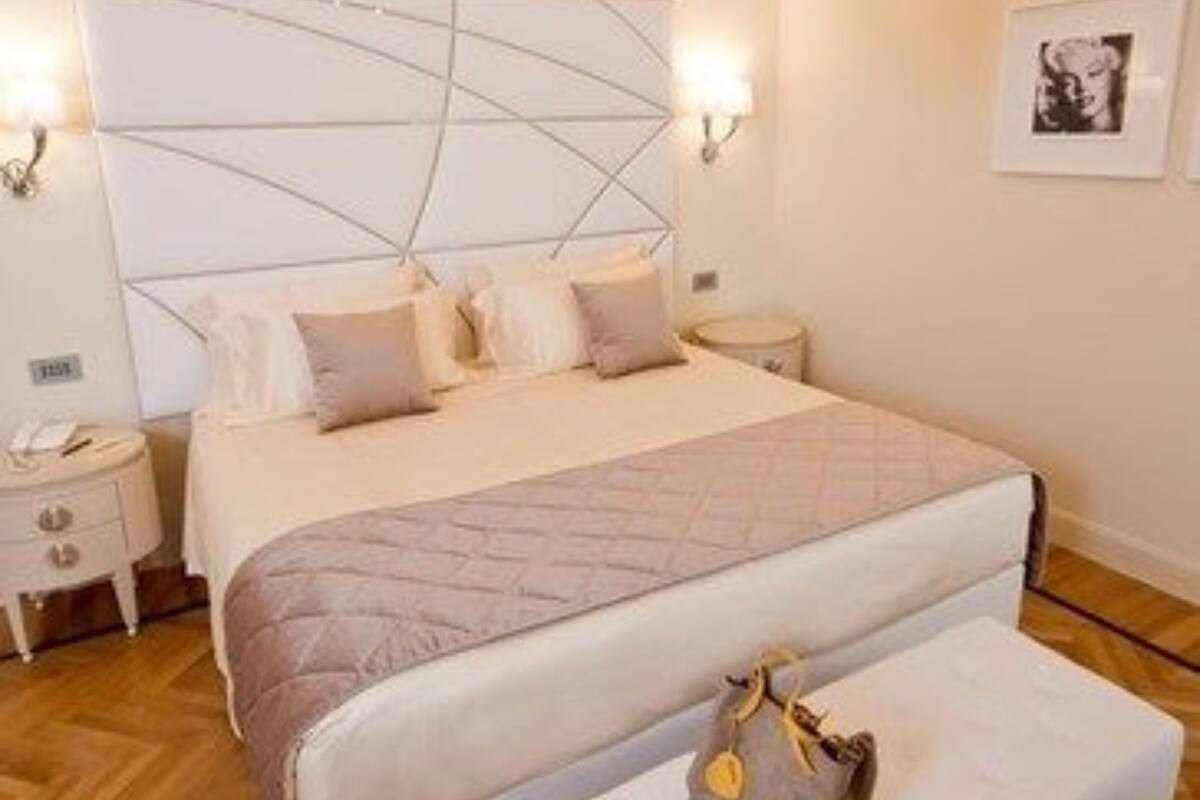 A bedroom with a large bed, a bedside table, a nightstand, a bench with a bag, and a chandelier.