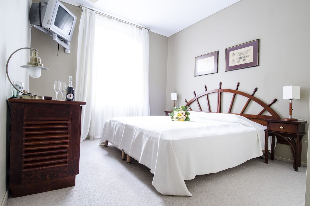 A hotel room with a bed, a wooden headboard, a desk with a lamp, a television mounted on the wall, and two framed pictures on the wall.