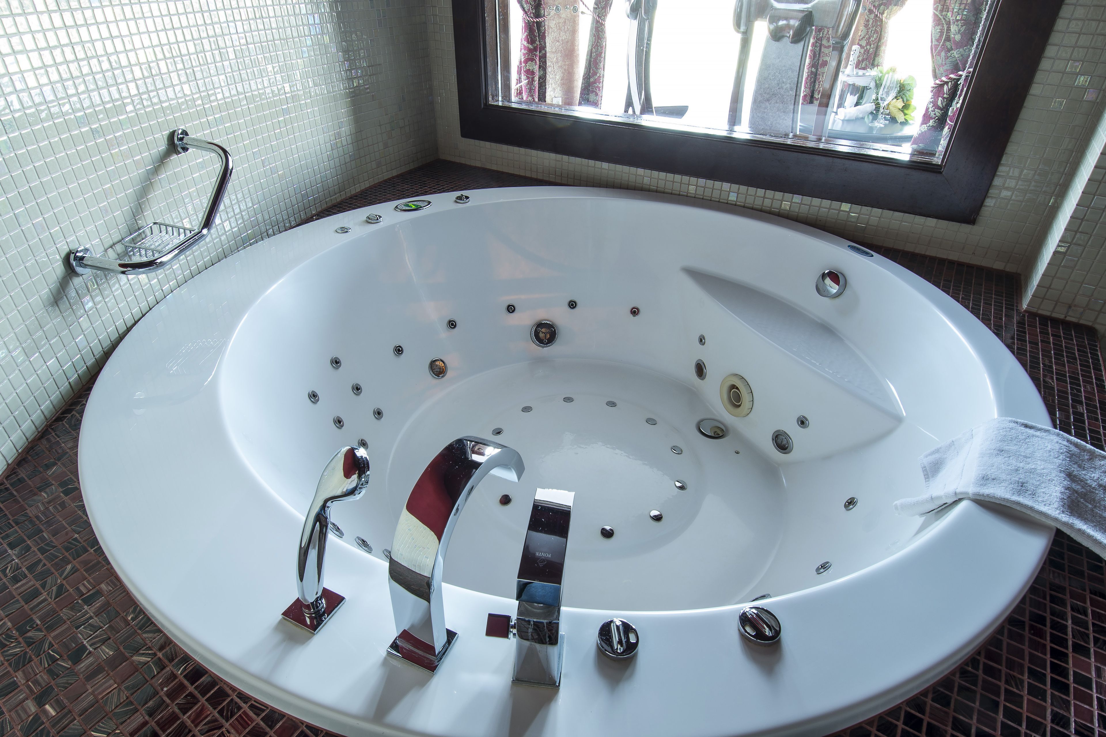 A large, round, white bathtub with multiple jets, chrome fixtures, and a glass shelf.