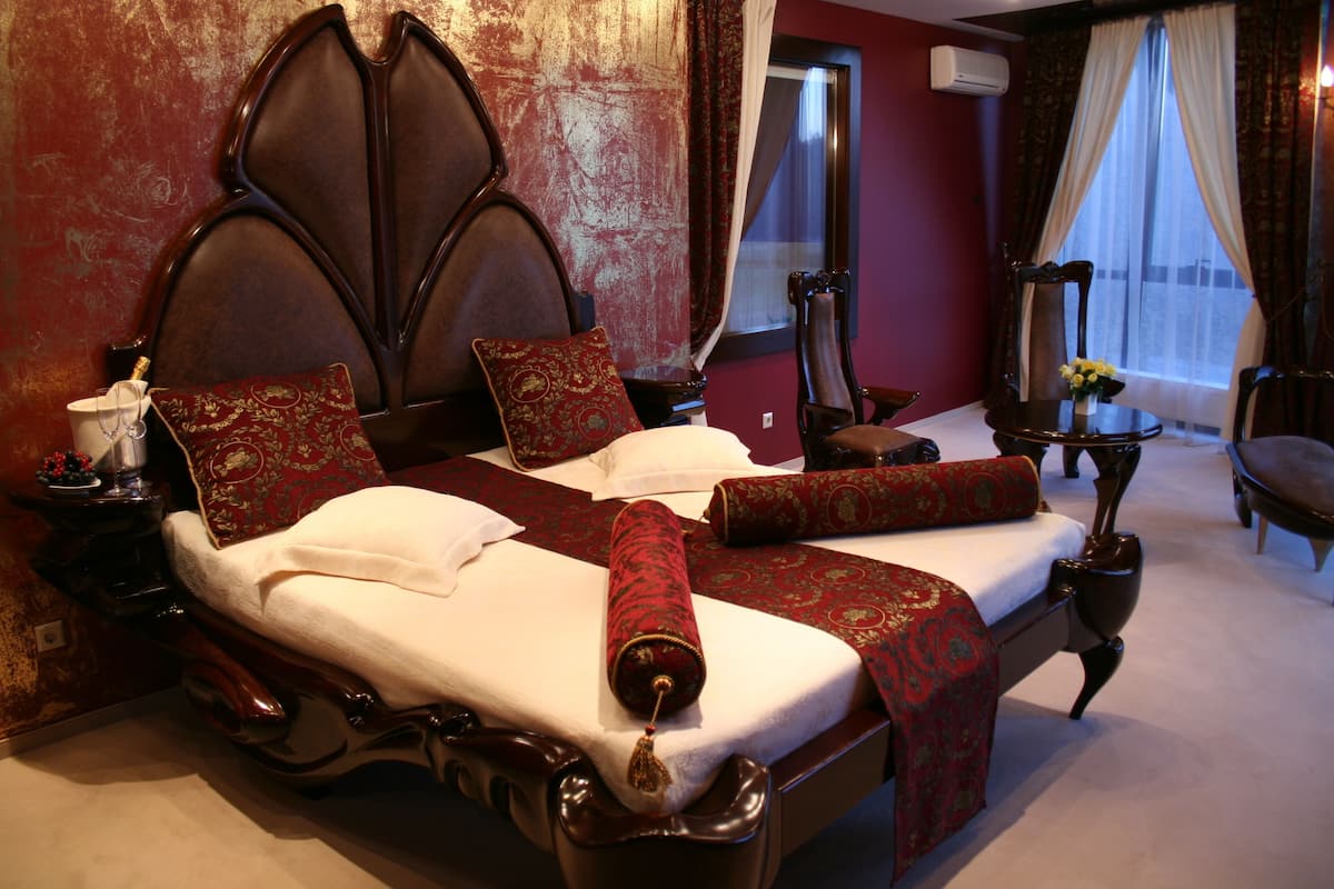 A large bed with ornate headboard, multiple pillows, and a red blanket. There is a small table with a vase of flowers and a chair beside the bed.