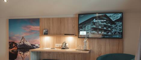 Una habitación de hotel moderna con un escritorio de madera, dos sillas color turquesa, una mesita redonda pequeña y una gran pantalla de televisión mostrando un complejo turístico de montaña.
