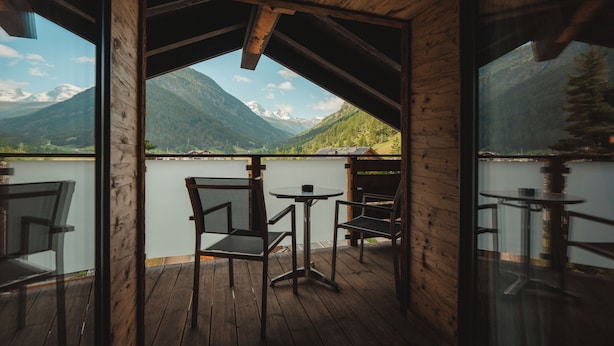 A wooden deck with a table and chairs, overlooking a lake and mountains.