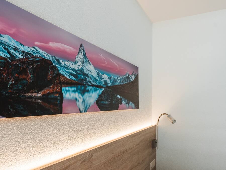 A bedroom with a wooden headboard, a wall-mounted lamp, and a large framed picture of a mountain landscape.