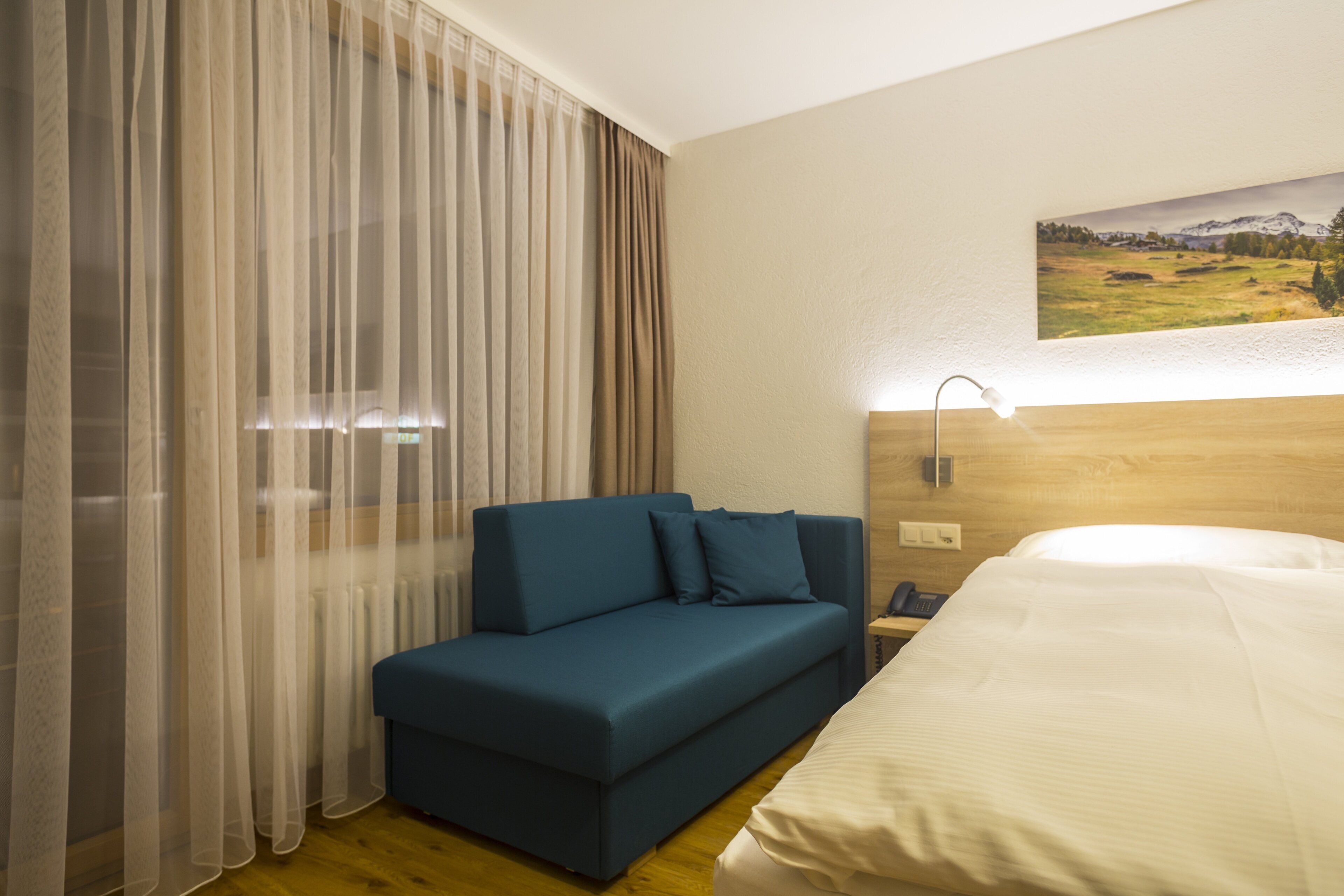 A hotel room with a bed, a blue sofa, a large window with sheer curtains, a painting on the wall, and a bedside lamp.