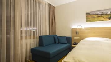 A hotel room with a bed, a blue sofa, a large window with sheer curtains, a painting on the wall, and a bedside lamp.
