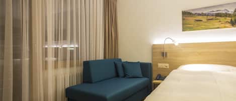 Habitación de hotel con cama, un sofá azul, un ventanal con cortinas translúcidas, un cuadro en la pared y una lámpara de noche.