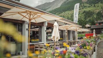 An outdoor seating area with umbrellas and tables, surrounded by flower beds, with a mountain backdrop.