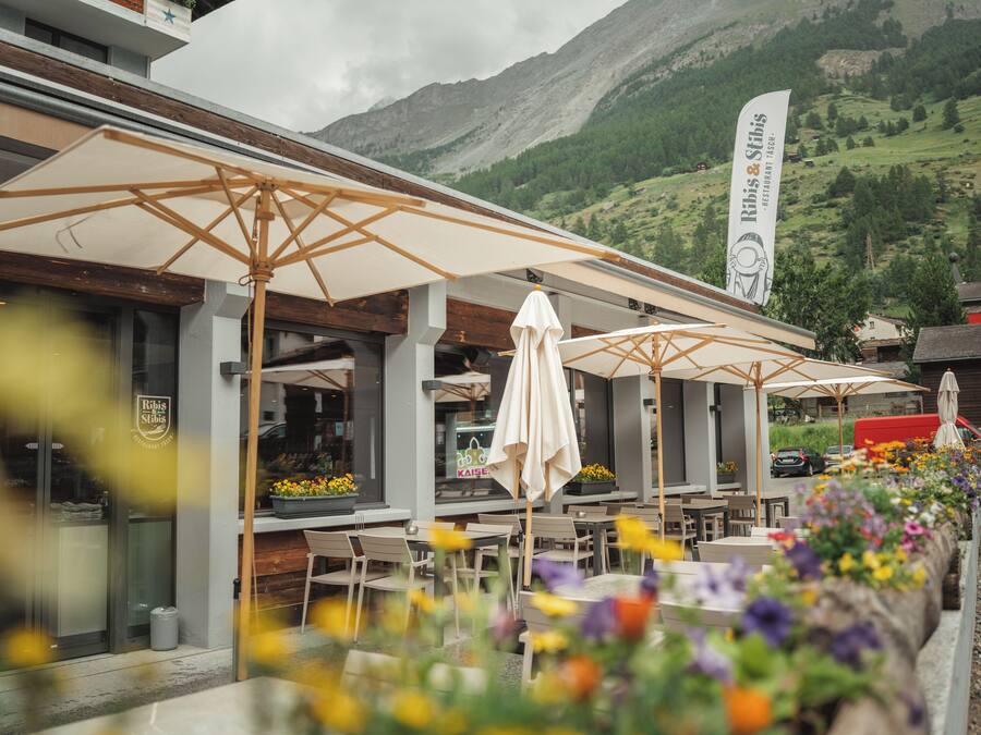 An outdoor seating area with umbrellas and tables, surrounded by flower beds, with a mountain backdrop.