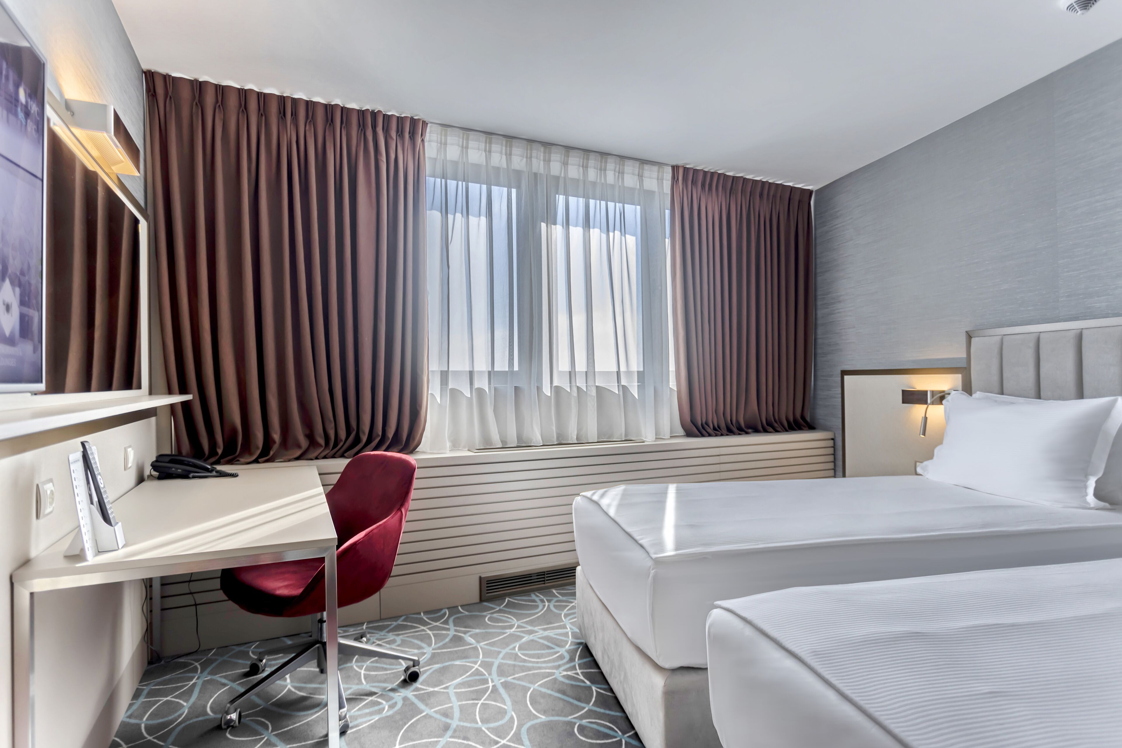 Una habitación de hotel con escritorio, una silla roja, una cama y un ventanal con cortinas.