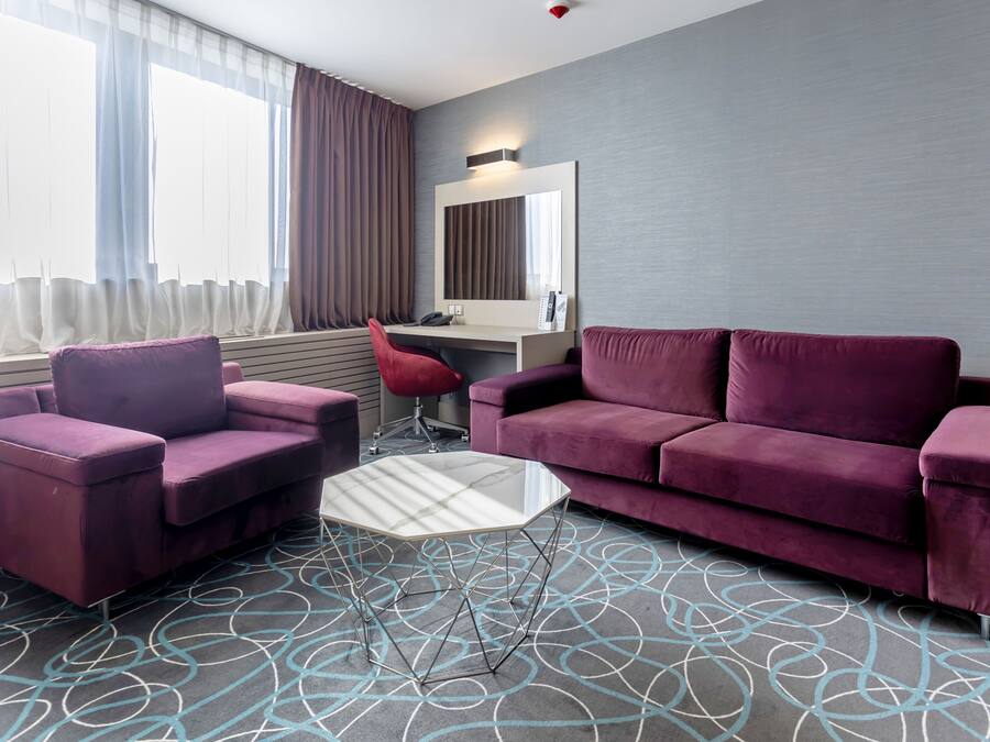 Ein modernes Hotelzimmer mit einem violetten Sofa, einem passenden Sessel, einem Glastisch und einem Schreibtisch mit Stuhl.