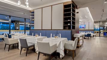 A modern restaurant with white tables and chairs, a blue cushioned bench, and a TV screen on the wall.