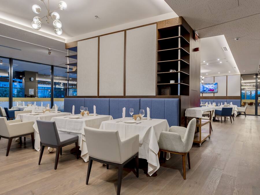 A modern restaurant with white tables and chairs, a blue cushioned bench, and a TV screen on the wall.
