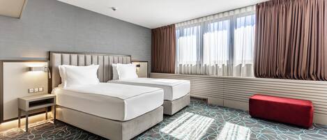 A modern hotel room with a large bed, two bedside tables, a red bench, and a large window with curtains.