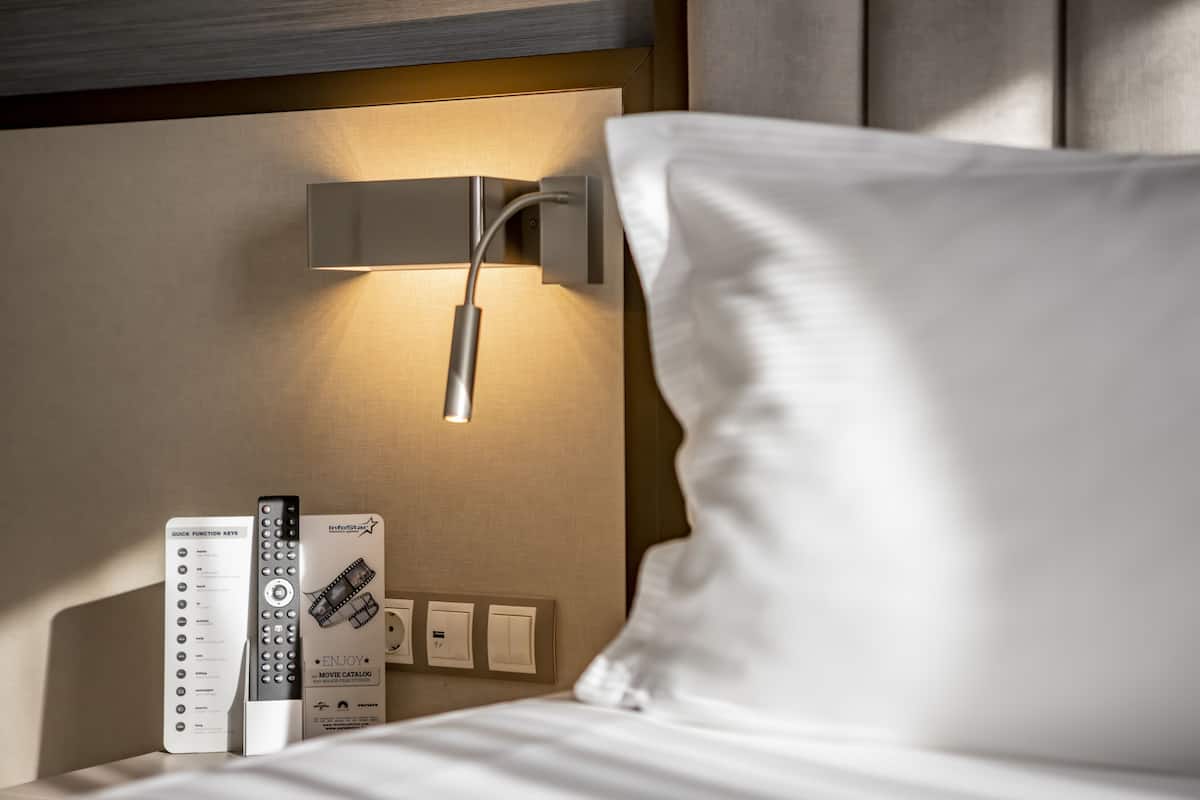 A hotel room with a bedside table, a remote control, a power outlet, and a wall-mounted light fixture.