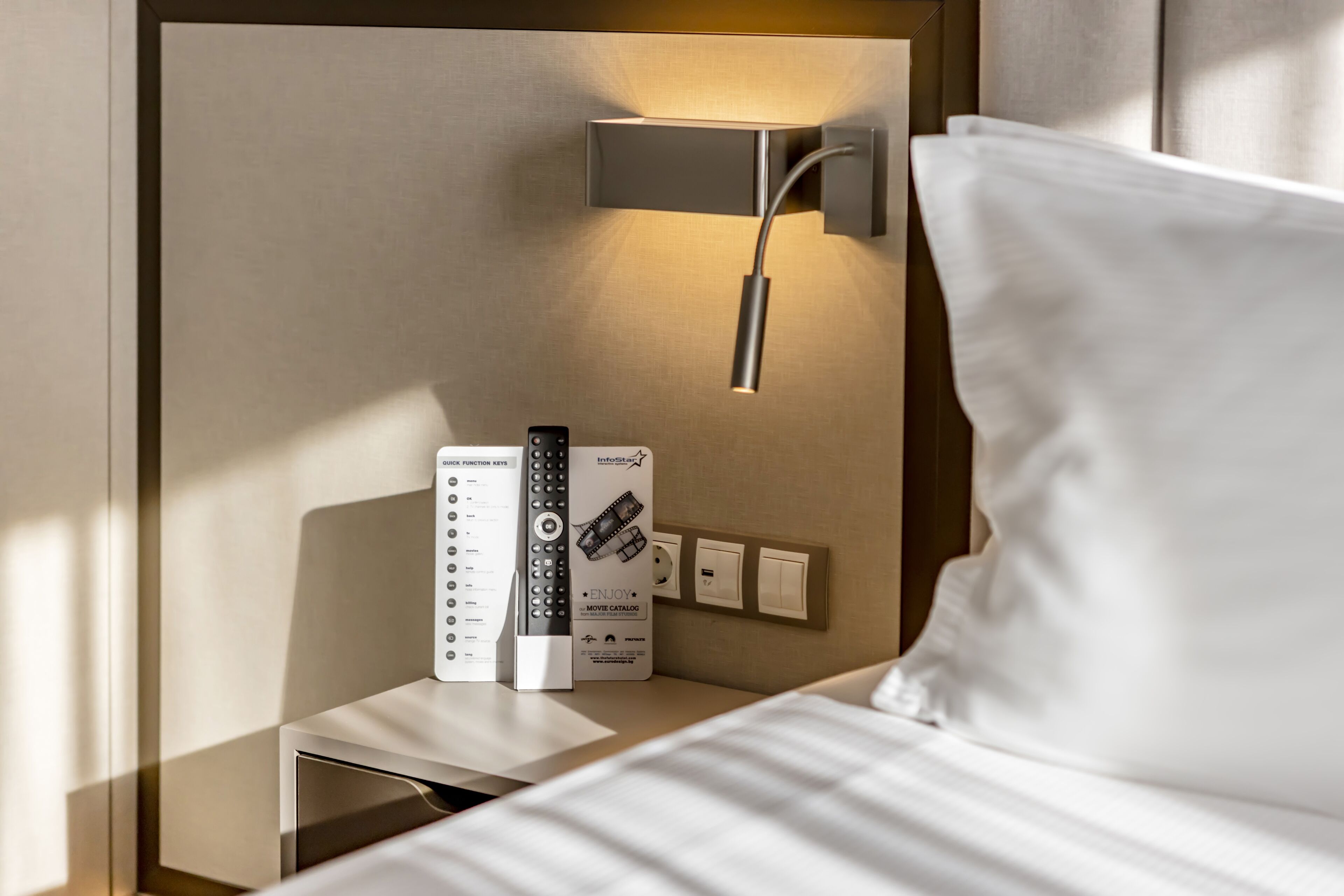 Ein Hotelzimmer mit Nachttisch, Fernbedienung, Steckdose und Wandleuchte.