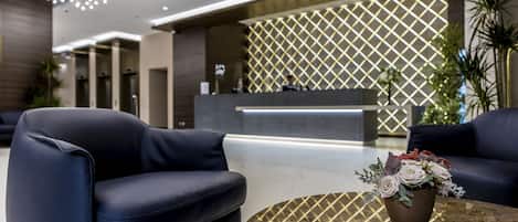 A modern reception area with a reception desk, two black leather sofas, a marble coffee table with a floral centerpiece, and a decorative wall with a geometric pattern.