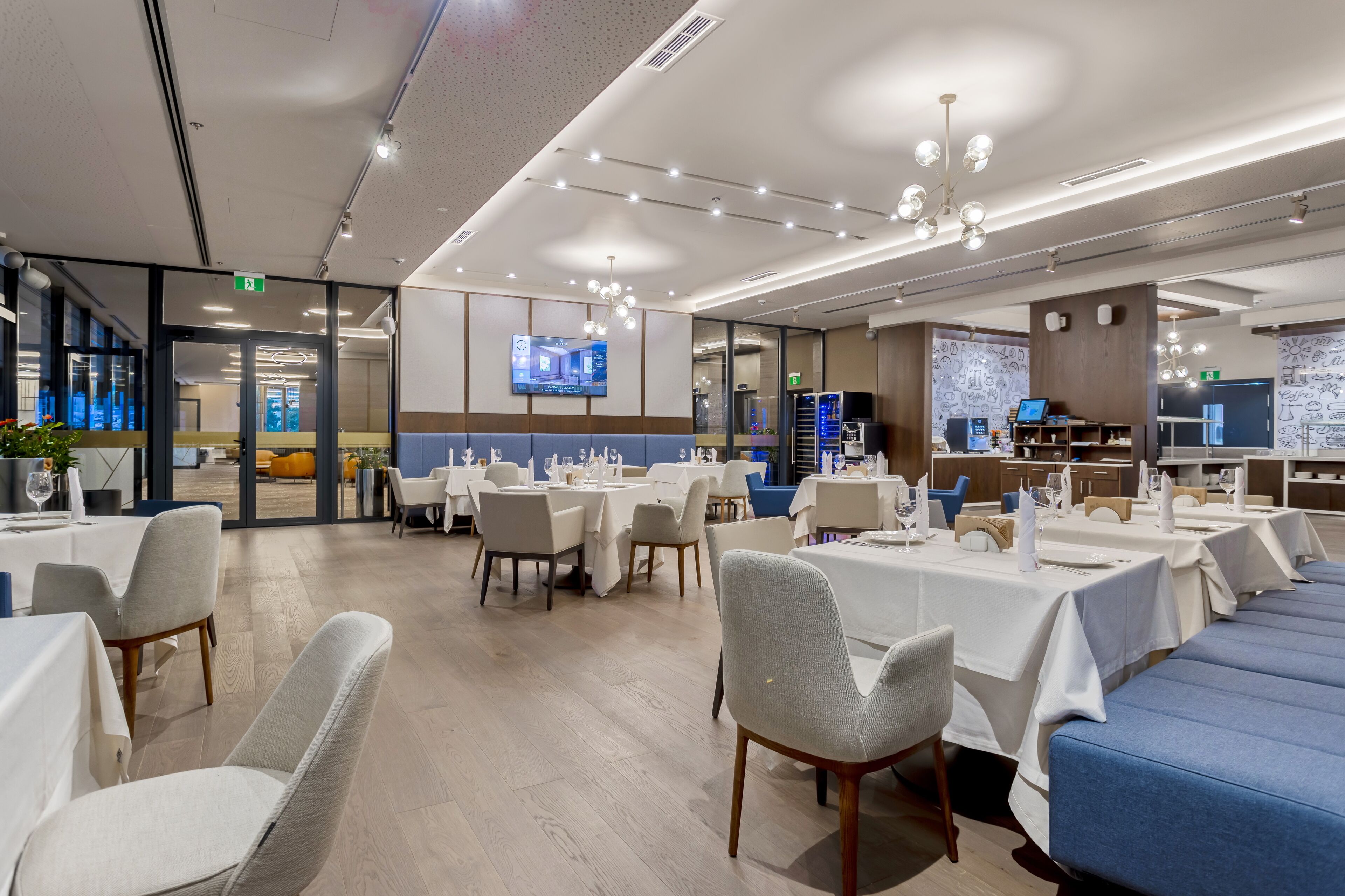 A modern dining area with tables and chairs, a television screen, and a kitchen area in the background.