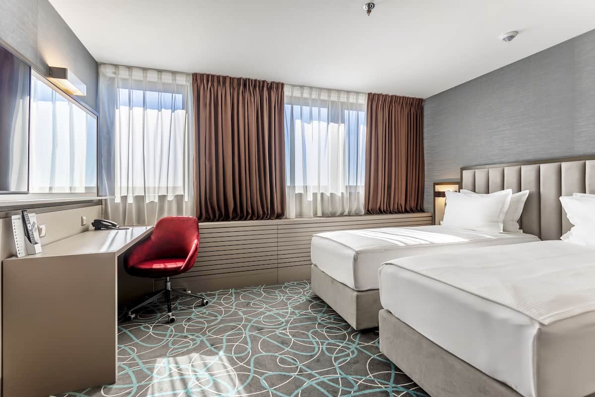 A hotel room with a desk, two beds, a red chair, and a patterned carpet.