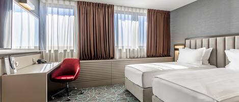 Habitación de hotel con escritorio, dos camas, una silla roja y ventanas grandes con cortinas.
