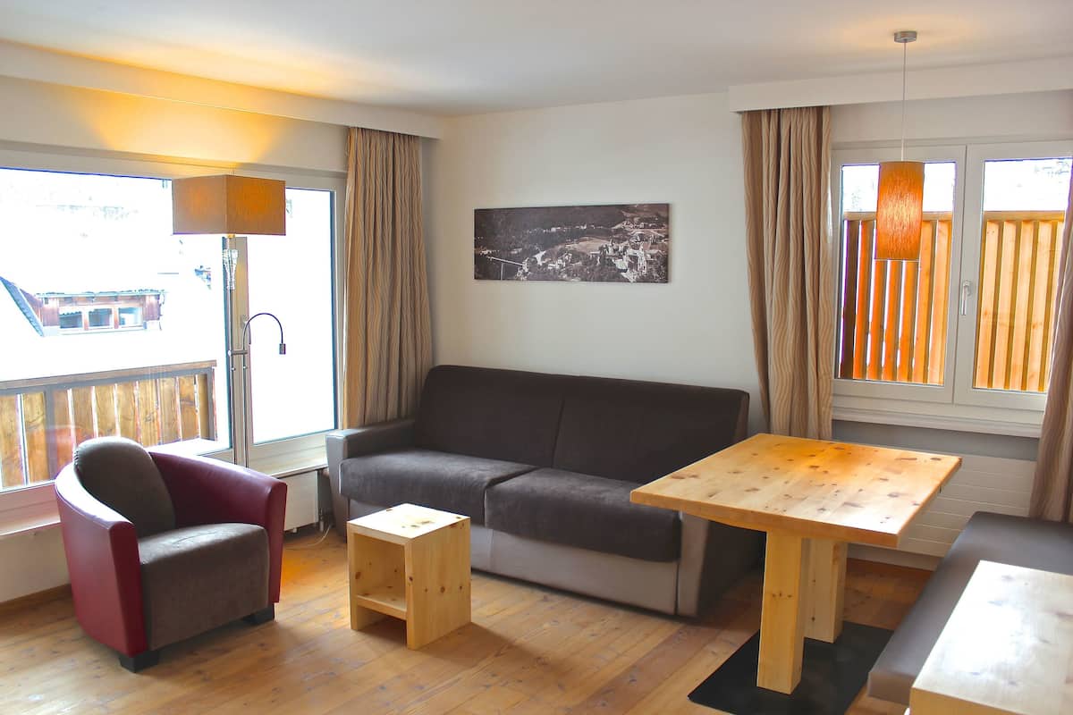 Una sala de estar con un sofá, un sillón rojo, una mesa de madera y vistas a un edificio a través de la ventana.