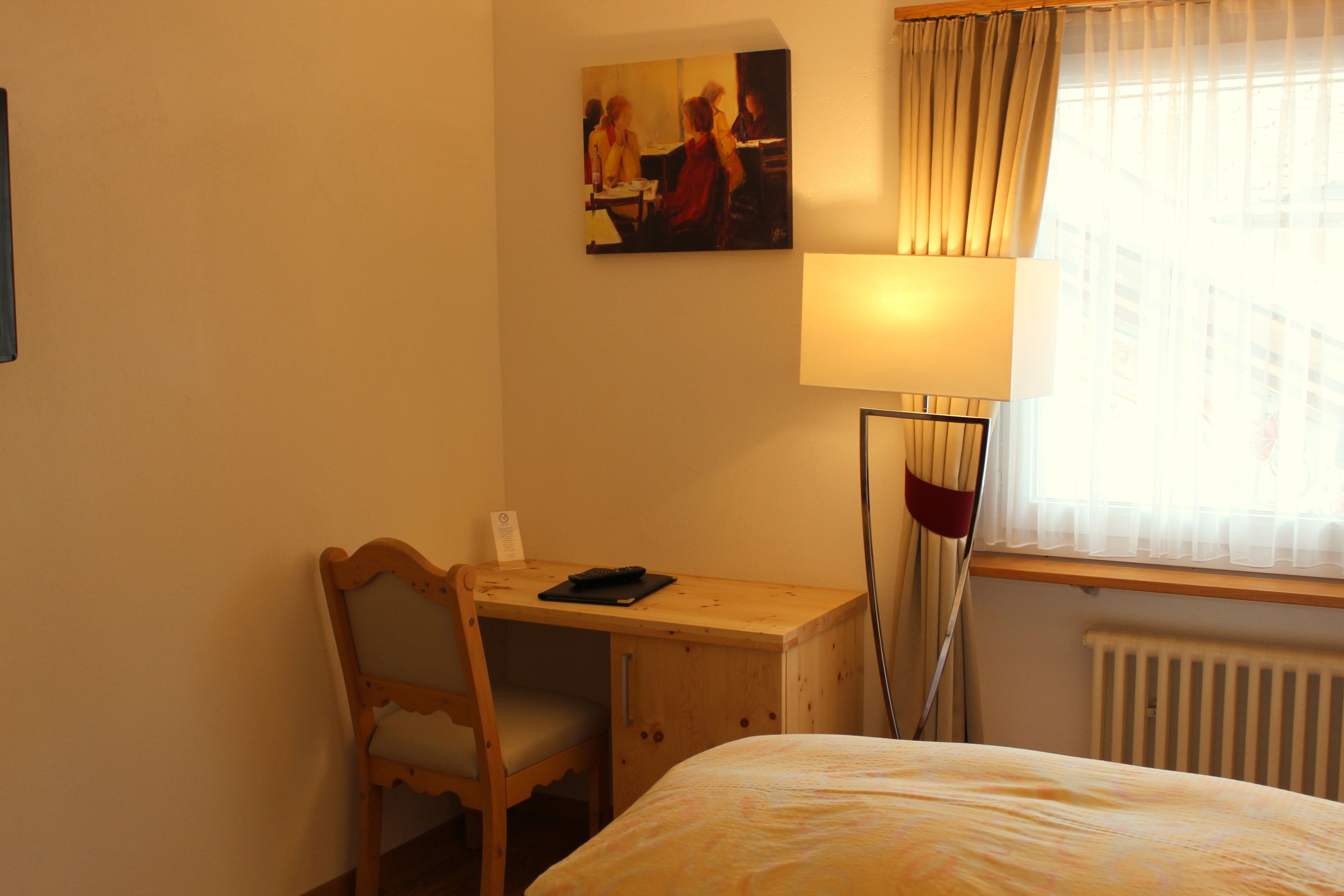 A hotel room with a wooden desk, a chair, a bed, a lamp, and a window with curtains.