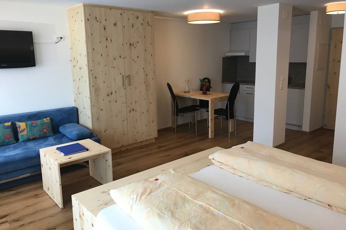 Una habitación con cama, sofá, una mesita, un armario de madera, una mesa de comedor con sillas y una zona de cocina.
