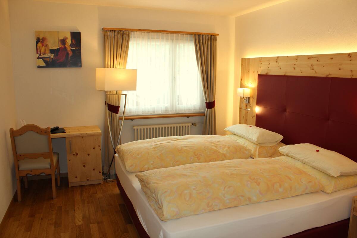 Habitación de hotel con dos camas, un escritorio, una silla, un ventanal con cortinas y un cabecero con luces.