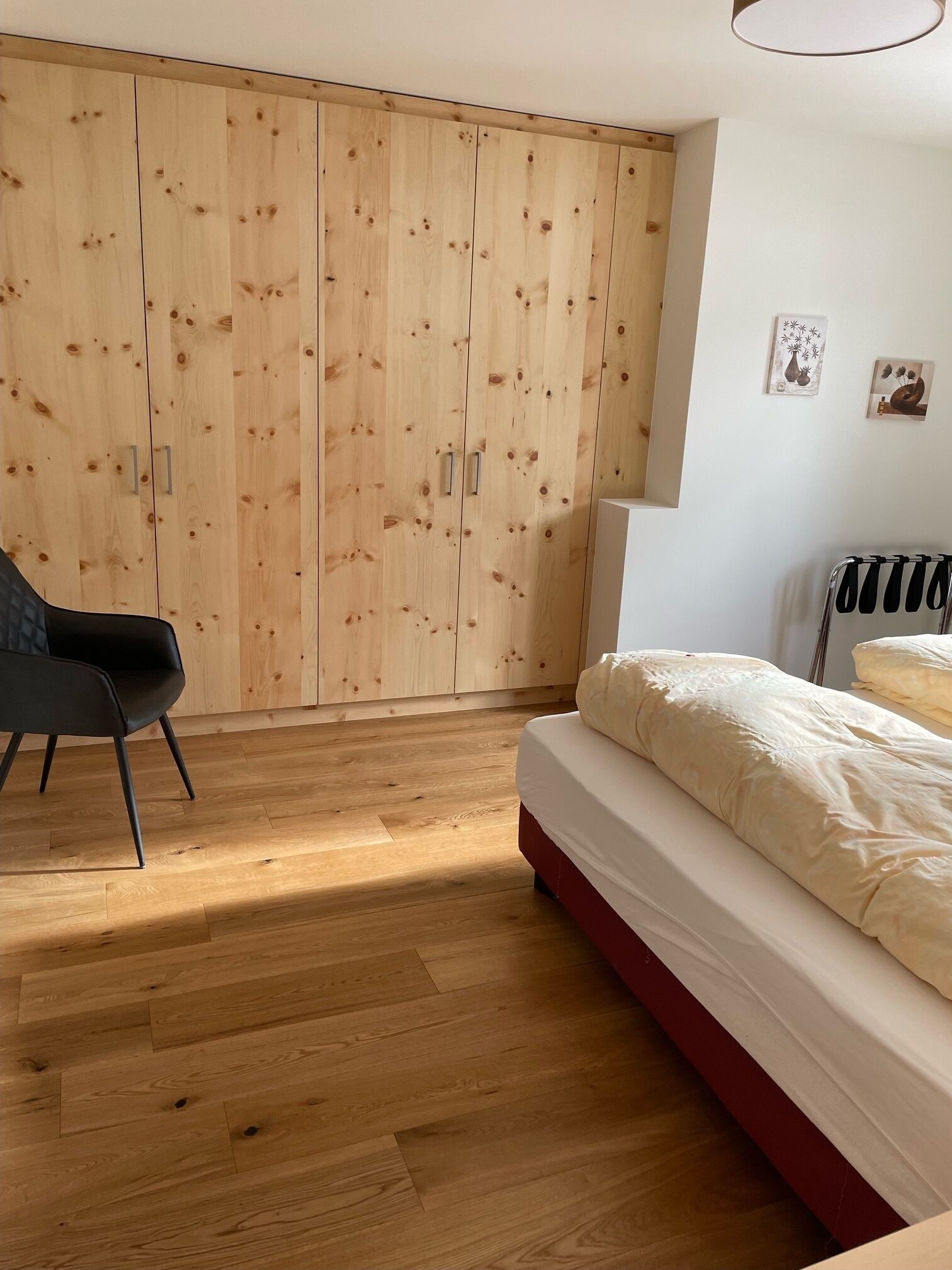 A bedroom with a wooden wardrobe, a bed with white bedding, a black chair, and a radiator.