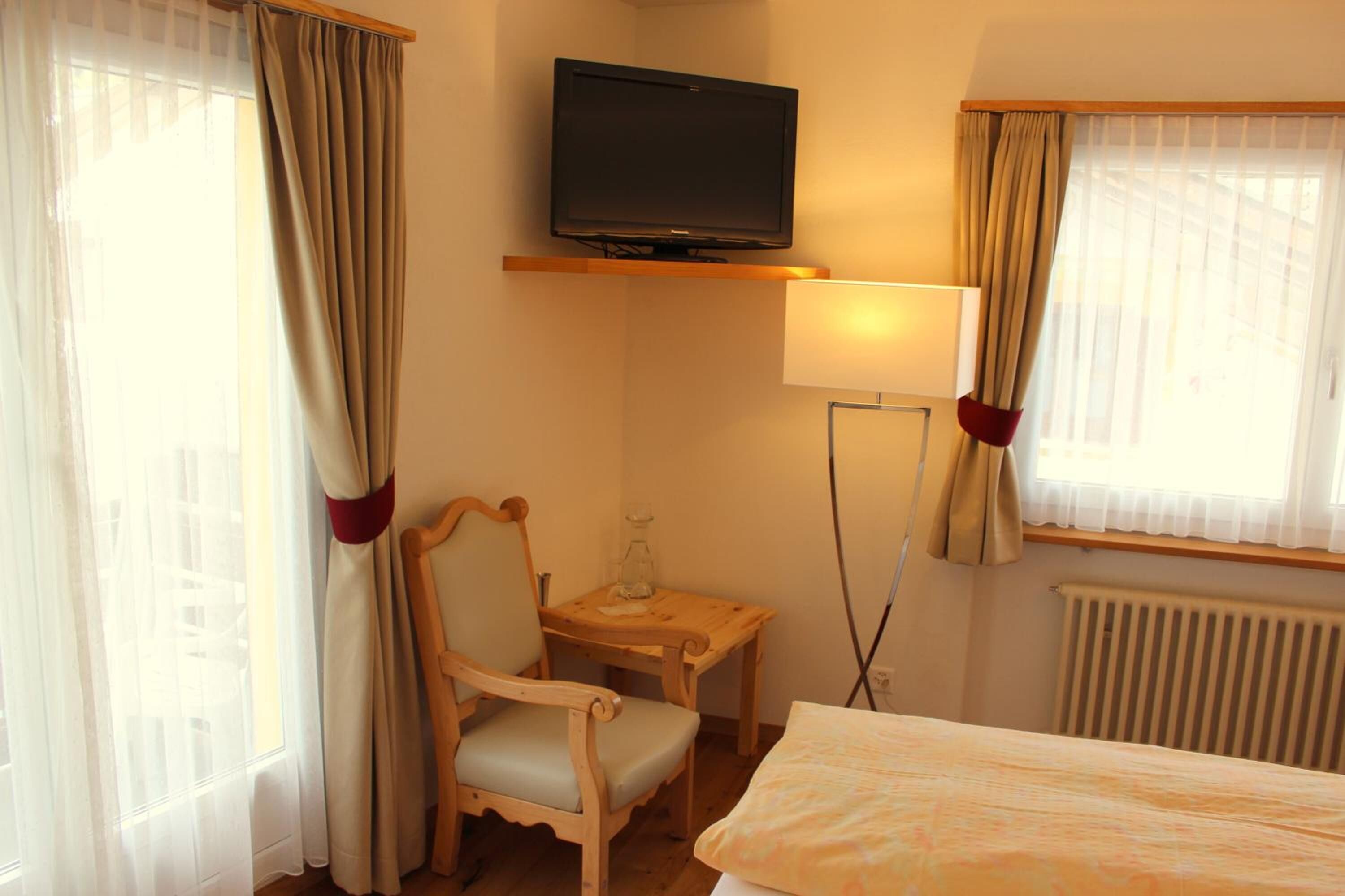 Una camera d'albergo con un letto, una TV a parete, un tavolino in legno, una sedia e una lampada.