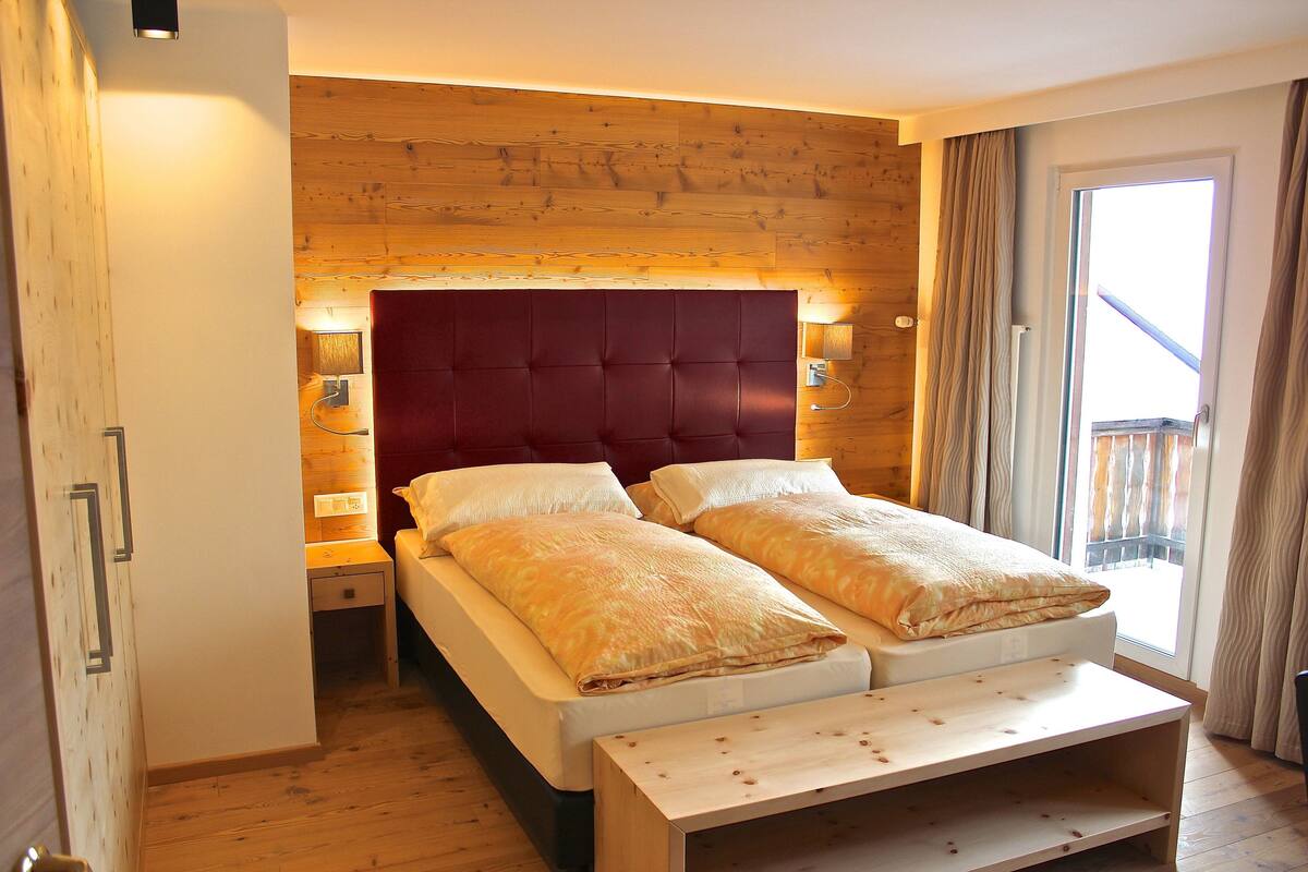 Una cama doble con cabecera de madera y dos lámparas de noche, un banco de madera al pie de la cama, y vistas a un balcón a través de una ventana con cortinas.
