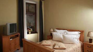A bedroom with a wooden bed, two bedside tables, a television on a wooden stand, a window with curtains, and a vase with flowers.